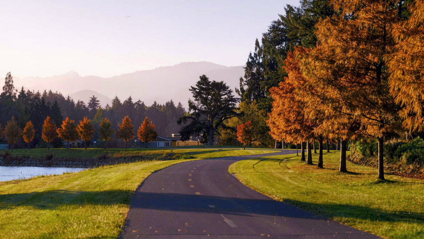 Wide paved community trail curving through open park space in an established Sammamish Washington plateau neighborhood with ornamental trees in autumn color, mature Douglas fir at the forest edge, and Cascade foothills softly visible in the background